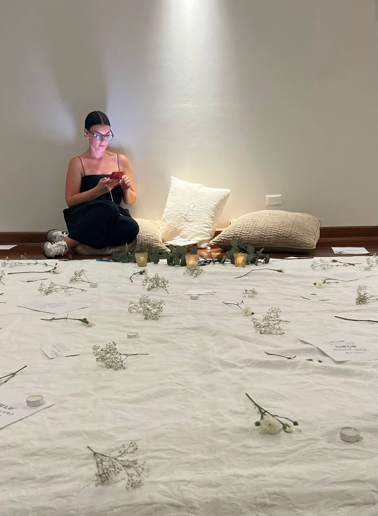 Mujer sentada en el suelo sobre un cojín con las piernas sentada. Frente a ella hay una manta en el suelo de color blanco sobre la cual hay varias flores blancas desperdigadas.
