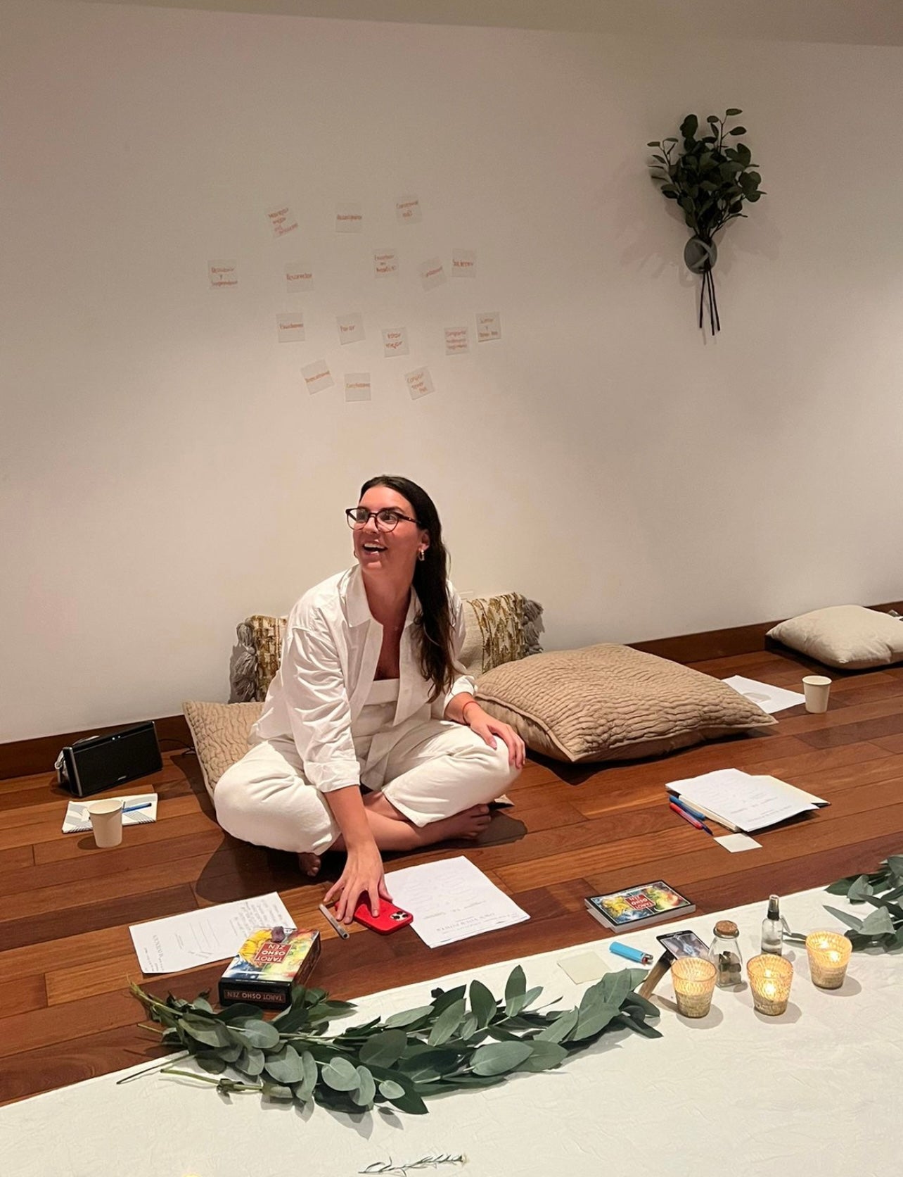 Mujer sonriente sentada sobre un cojín en el suelo con las piernas cruzadas. Frente a ella hay velas, papeles y ramas de eucalipto. 