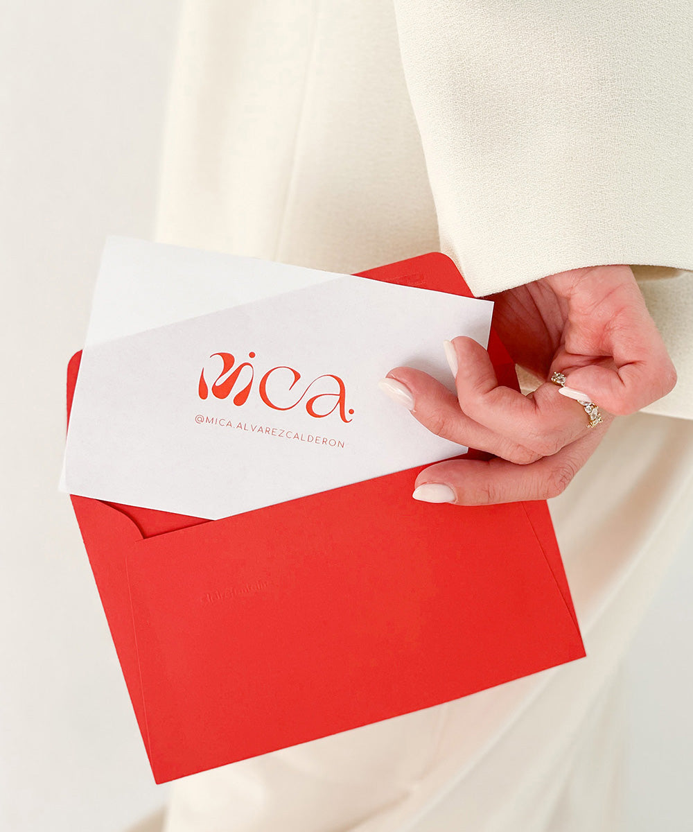 Acercamiento de una mano sosteniendo un sobre de color rojo. Del sobre se asoma un papel con el logo de Mica impreso en color rojo. 