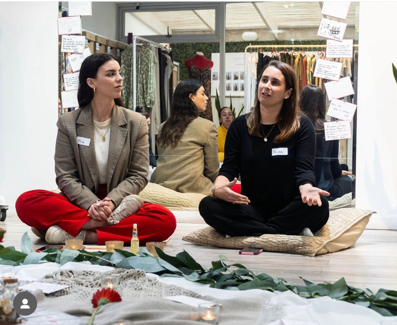Dos mujeres charlando sentadas con las pierna cruzadas sobre el suelo. Detrás de ellas hay un espejo en el cual se ven reflejados múltiples racks de ropa. 